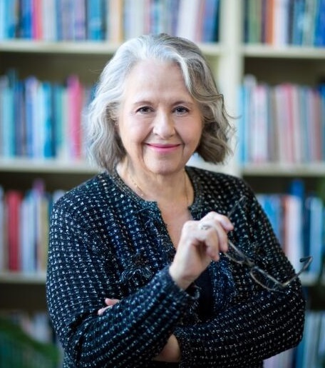 Head and shoulder picture of an older woman with grey-white shoulder-length hair standing in front of a shelf of books, holding glasses