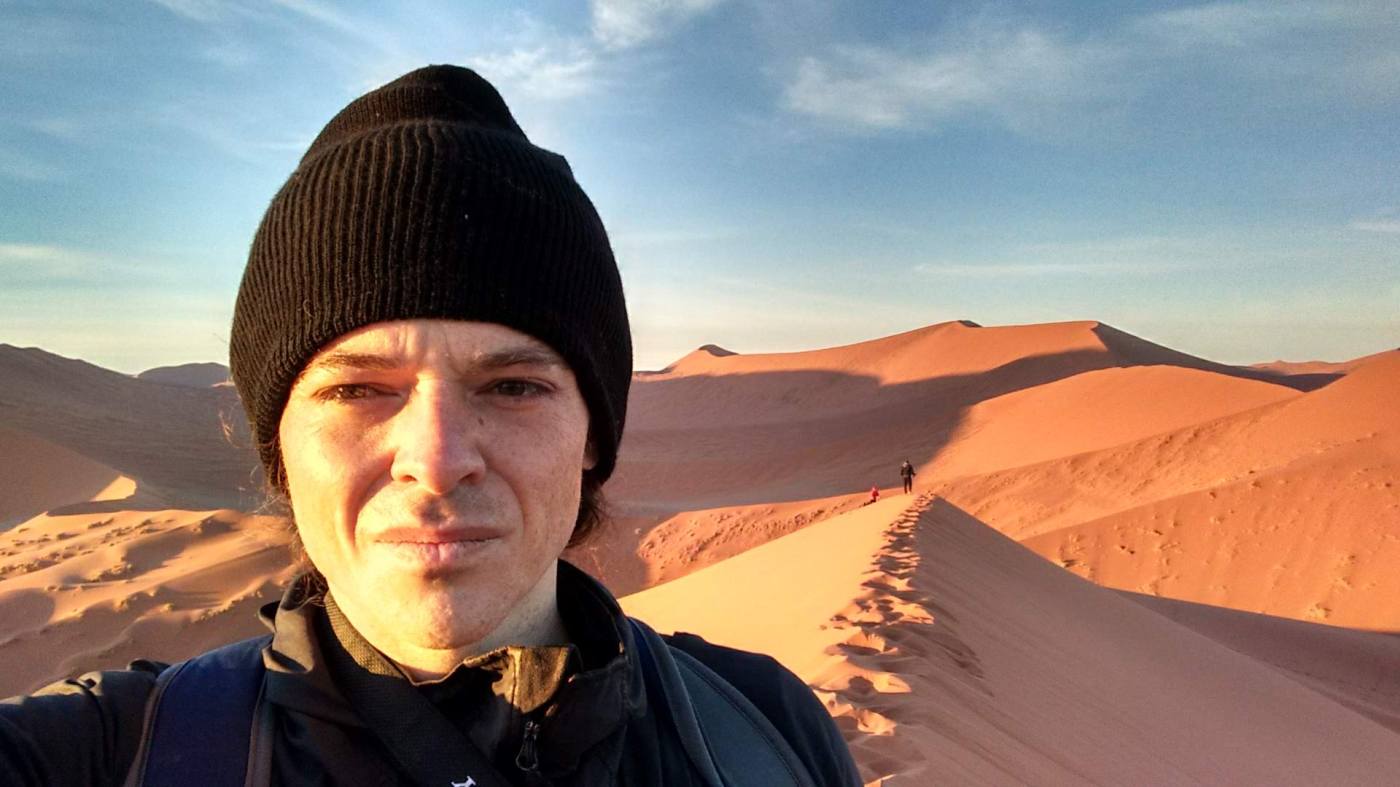 Author Jesse O'Reilly-Conlin wearing a black toque, black shirt and a backpack standing in front of mountains of red sand